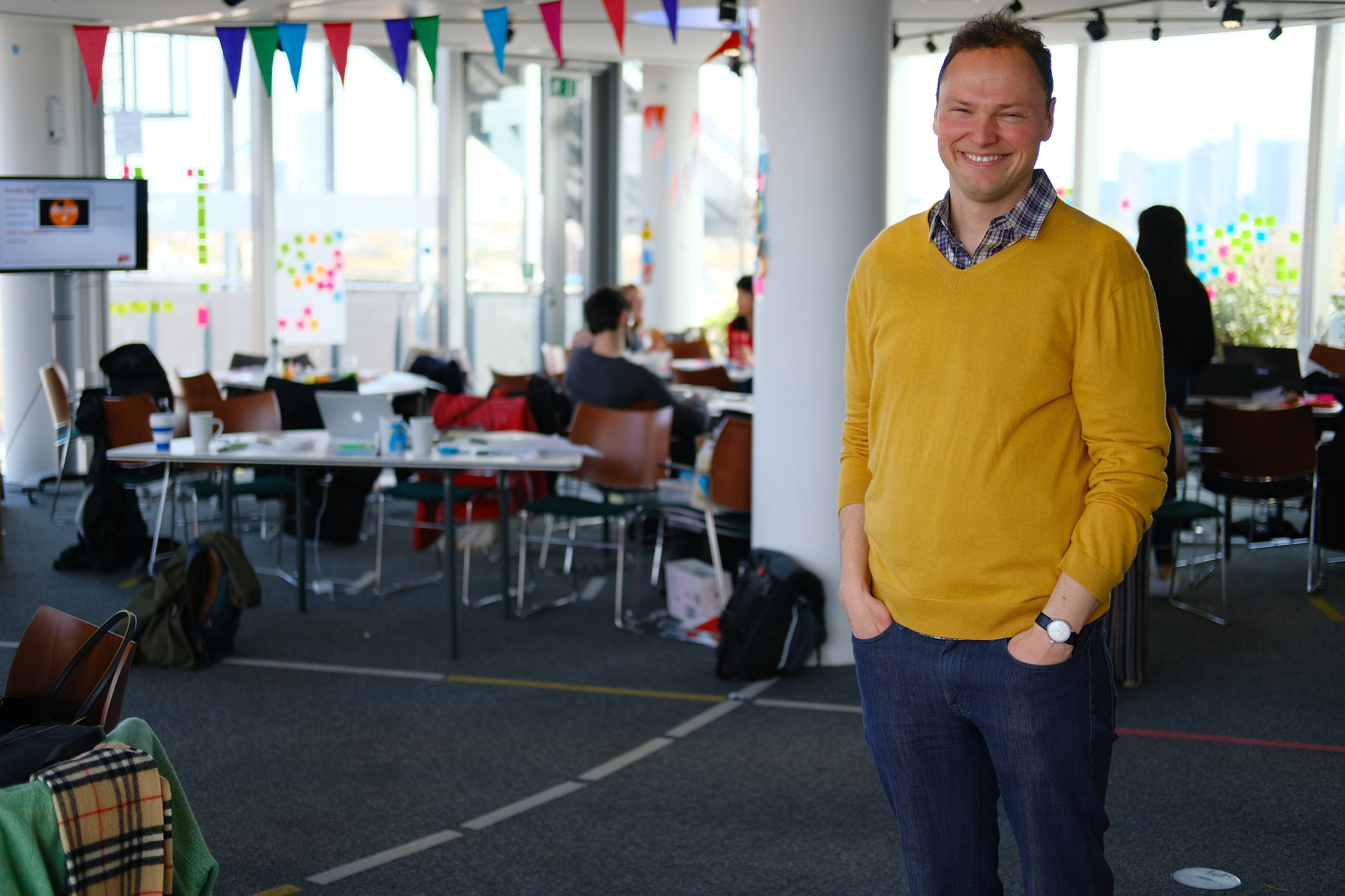 Andreas Conradi smiling in a workshop space, with sticky notes and a design studio setup in the background.