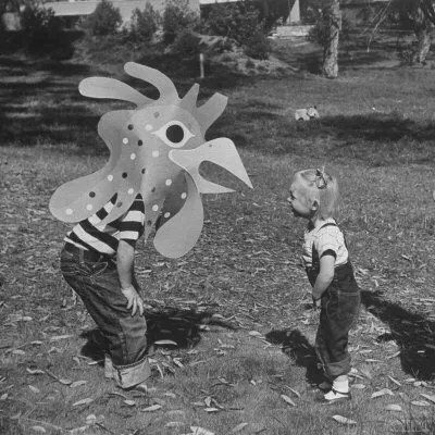 Vintage black-and-white photo of a child in a colourful paper-bird mask facing a small child in dungarees on a lawn.