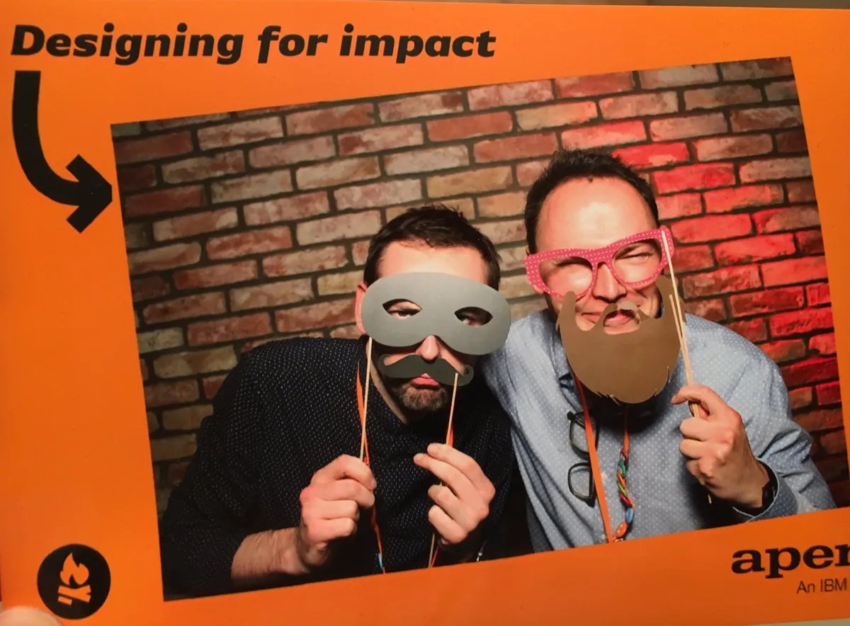 Photo-booth print reading &lsquo;Designing for impact&rsquo;, with two colleagues posing in paper masks in front of a brick wall, from an Apperio/IBM event.