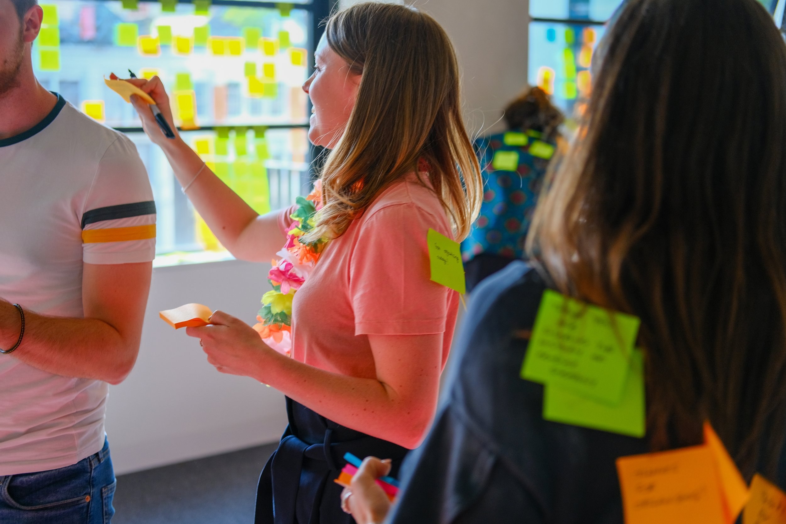 A colleague at a product away day sticking notes to a teammate's t-shirt, in front of windows covered in sticky notes.
