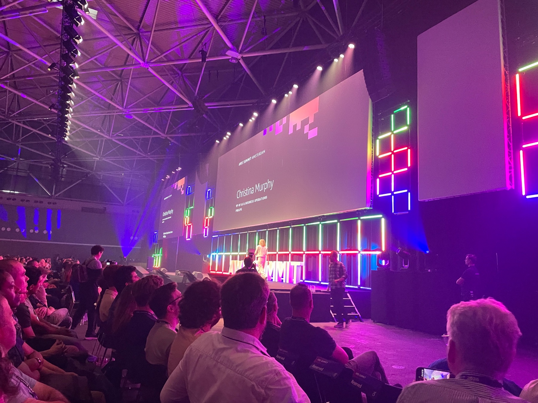 A keynote speaker on stage in front of a large audience at the AWS AI Summit, under colourful stage lighting.