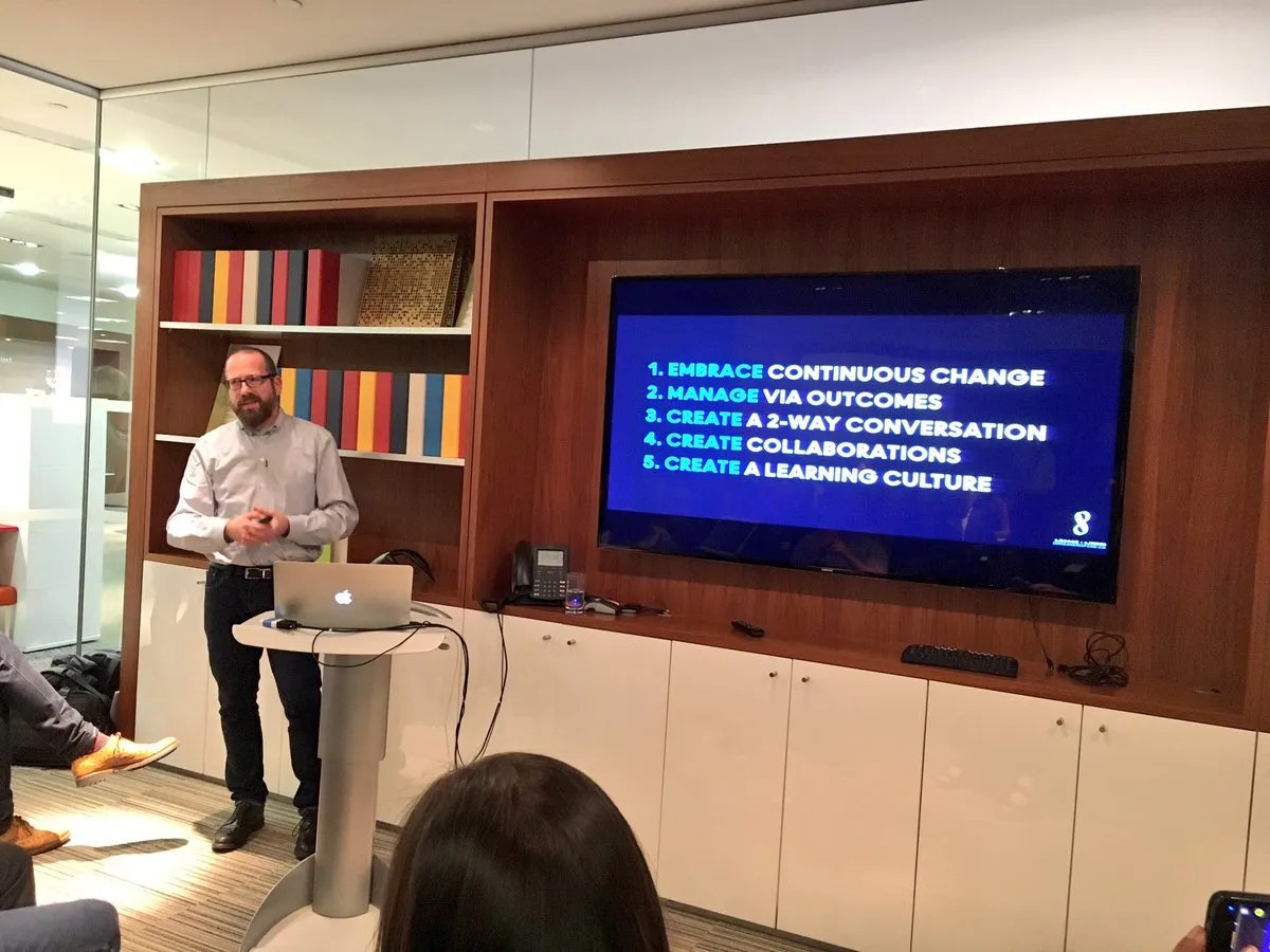 Speaker presenting a slide listing five principles: embrace continuous change, manage via outcomes, create a 2-way conversation, create collaborations, create a learning culture.
