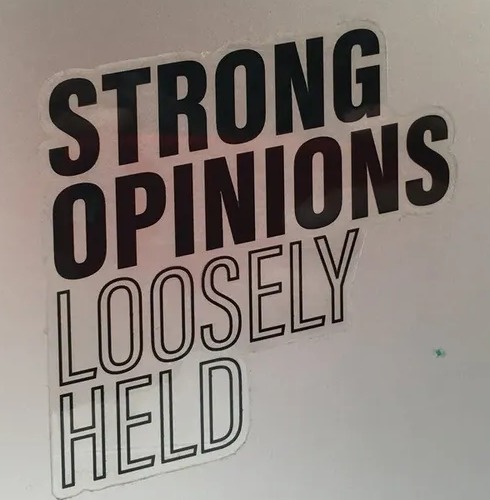 A physical sticker reading &lsquo;Strong opinions, loosely held&rsquo; stuck on a laptop lid.